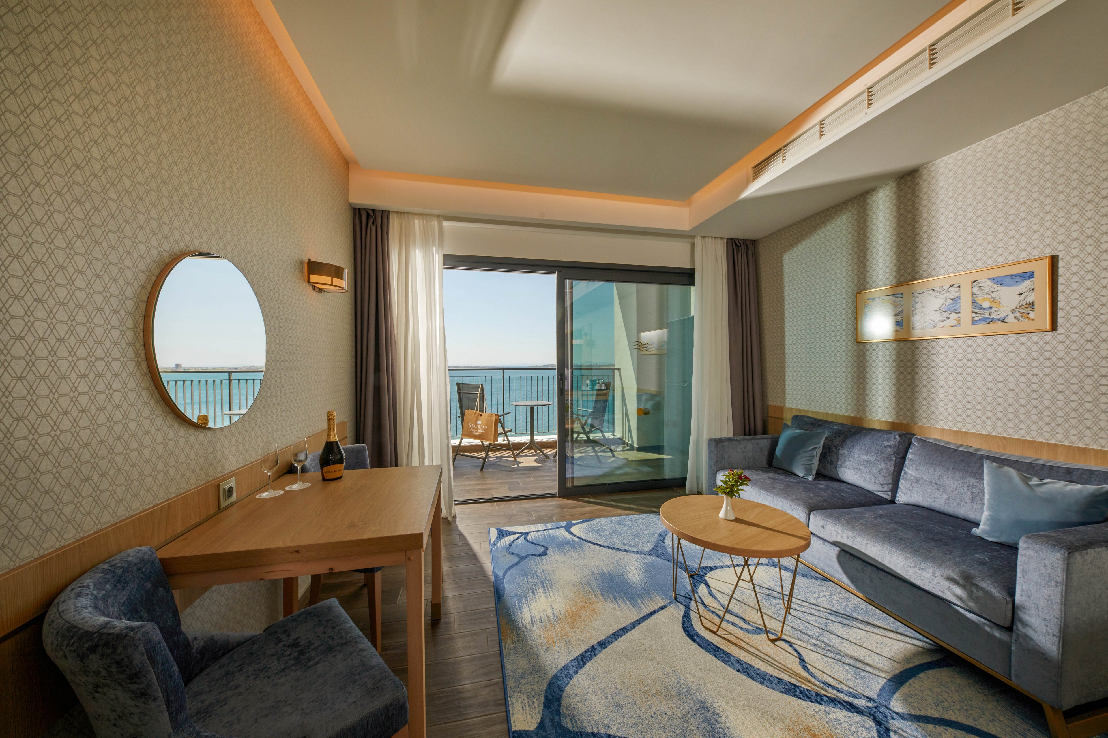 A contemporary hotel room features a cozy seating area with a blue sofa and patterned rug. A wooden desk with a round mirror and a bottle with glasses sits near the wall. Large sliding glass doors open to a balcony with chairs overlooking the sea. Soft lighting and neutral tones create a calm, relaxing atmosphere ideal for vacation or business travel.
