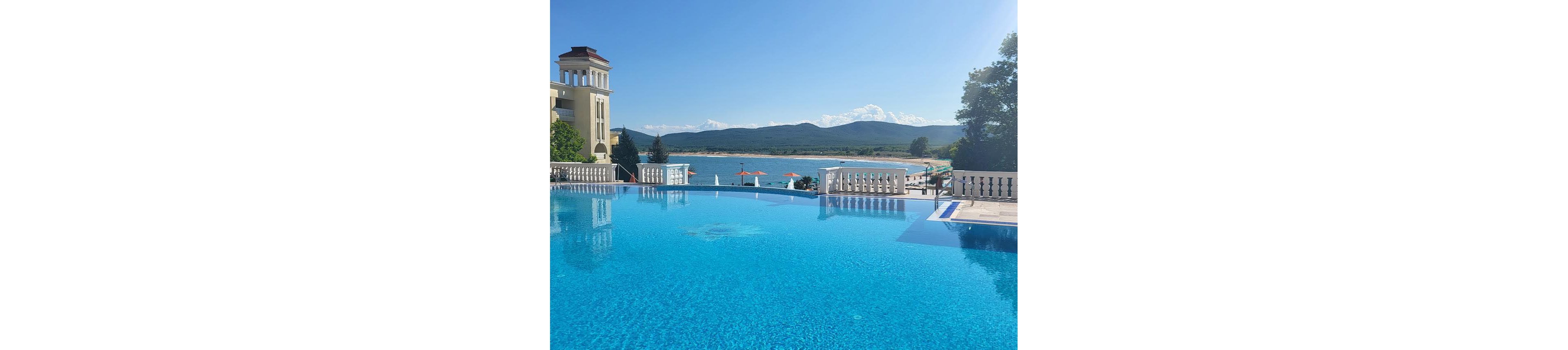 A clear blue infinity pool stretches toward a scenic lake and distant mountains under a bright, cloud-dotted sky. Elegant balustrades and a classical-style building frame the poolside area, suggesting a high-end resort setting. Sun loungers and orange parasols line the waterfront, adding a relaxed vacation atmosphere. The overall scene conveys calm, leisure, and upscale summer travel.