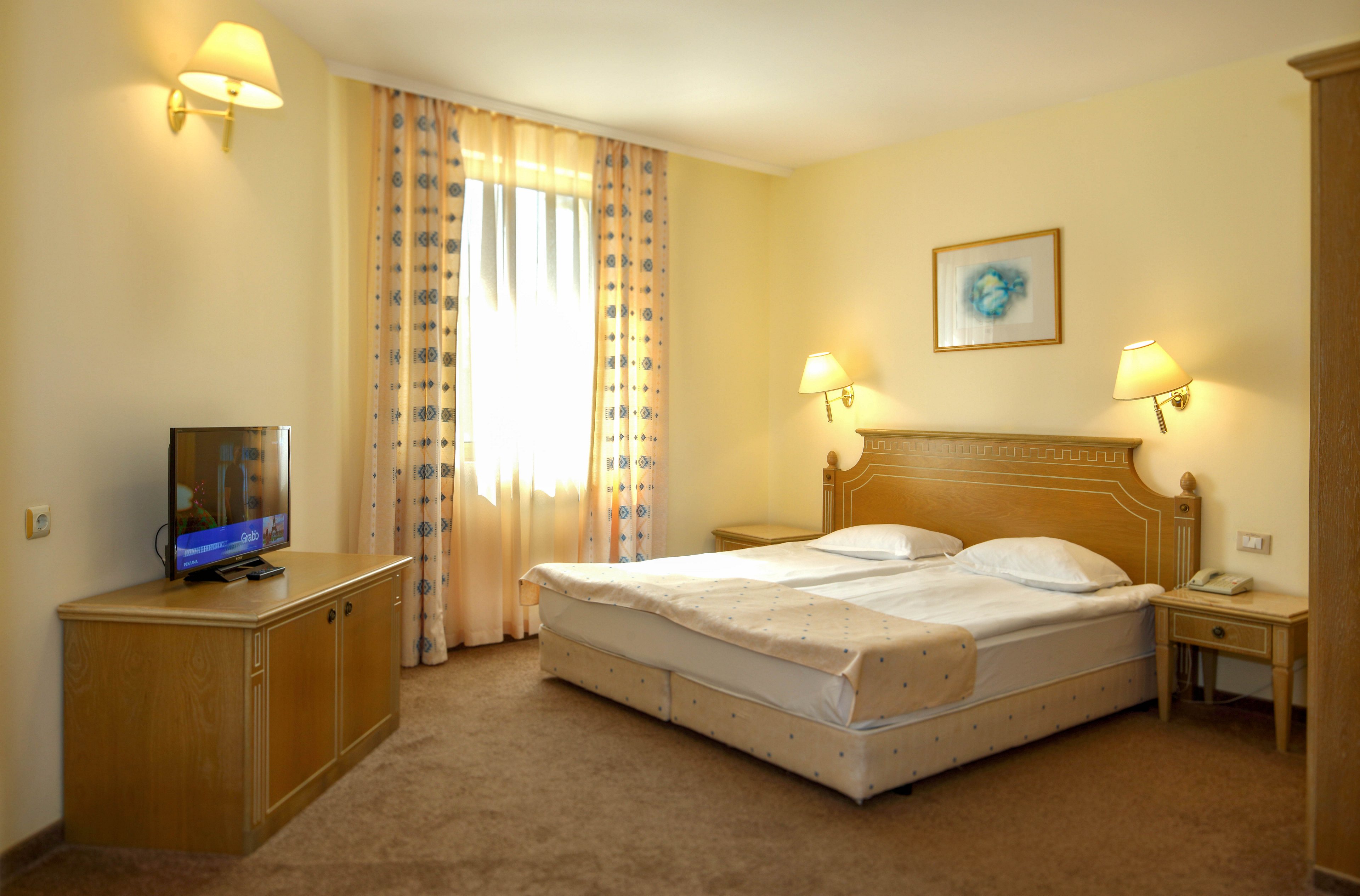 A tidy hotel bedroom features a large double bed with white linens and a light beige bed runner. The room includes wooden furniture, such as bedside tables, a TV stand with a flat-screen television, and a small desk with a telephone. Soft yellow walls, wall-mounted lamps, and patterned curtains framing a tall window create a warm, inviting atmosphere. A framed artwork above the bed adds a decorative touch to the simple, comfortable space.