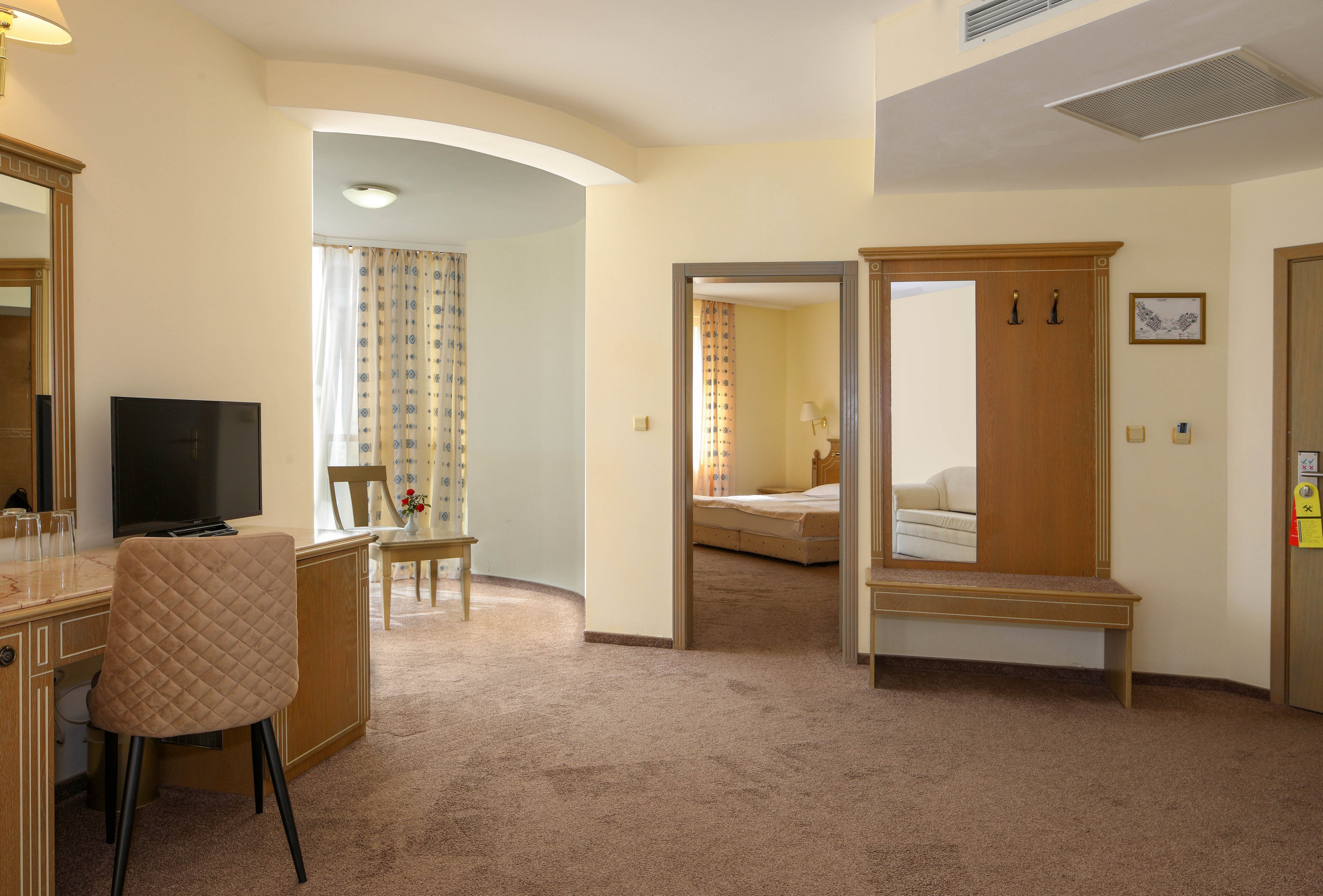 Interior view of a spacious hotel room featuring a desk with a chair and television in the foreground. An open doorway leads to a separate bedroom area with a neatly made bed and matching curtains. Warm beige walls and carpet create a soft, inviting atmosphere, complemented by wooden furniture and soft lighting. A large mirror, coat hooks, and small decorative details add functionality and comfort to the space.