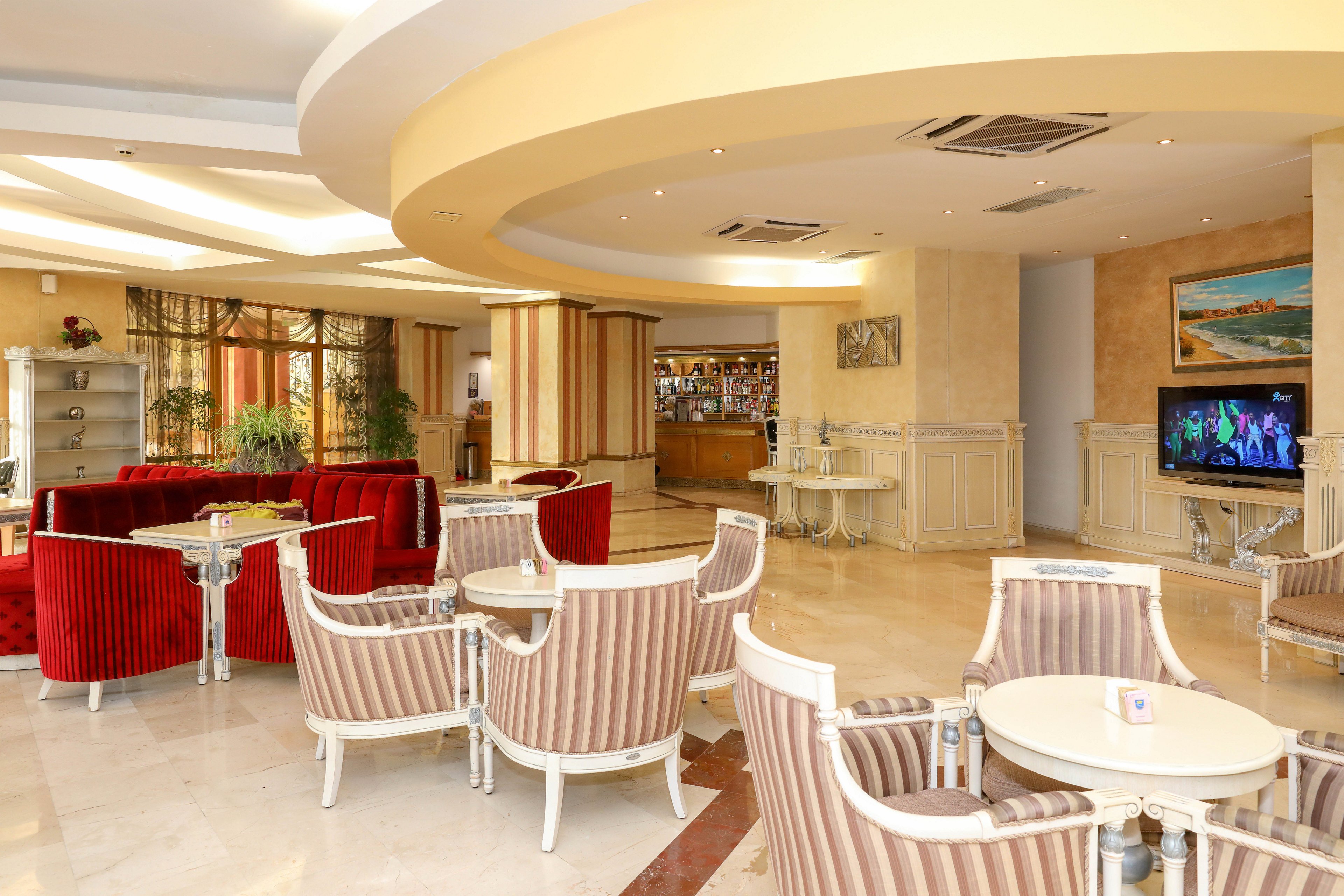 Bright hotel lobby lounge featuring striped armchairs and round white tables arranged for conversation. Rich red sofas add contrast against cream-colored walls and marble flooring. A bar area is visible in the background, along with decorative plants, artwork, and a television screen displaying colorful imagery. The space is well-lit with recessed ceiling lights and curved architectural details, creating a welcoming atmosphere.