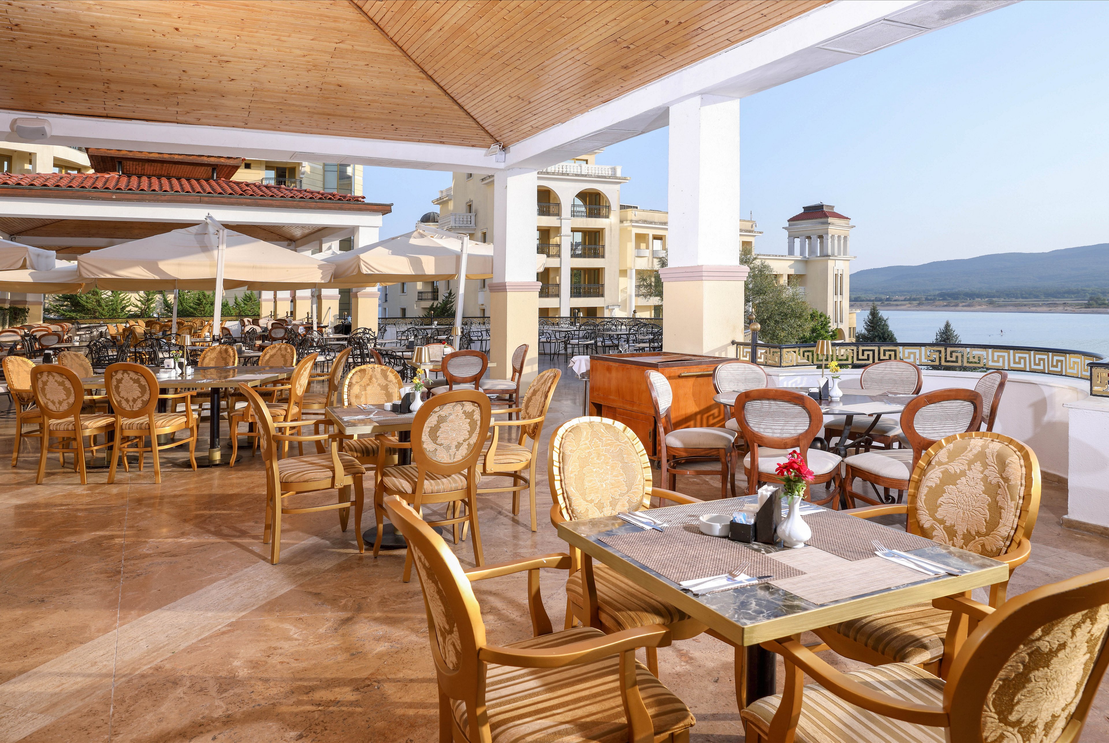 An open-air hotel terrace restaurant features neatly arranged wooden tables and chairs overlooking a calm waterfront. Sun umbrellas provide shade across the tiled patio, with folded napkins and cutlery set on each table. Mediterranean-style buildings and a distant shoreline create a relaxed vacation atmosphere. The scene is bright and inviting, suggesting a peaceful outdoor dining experience.