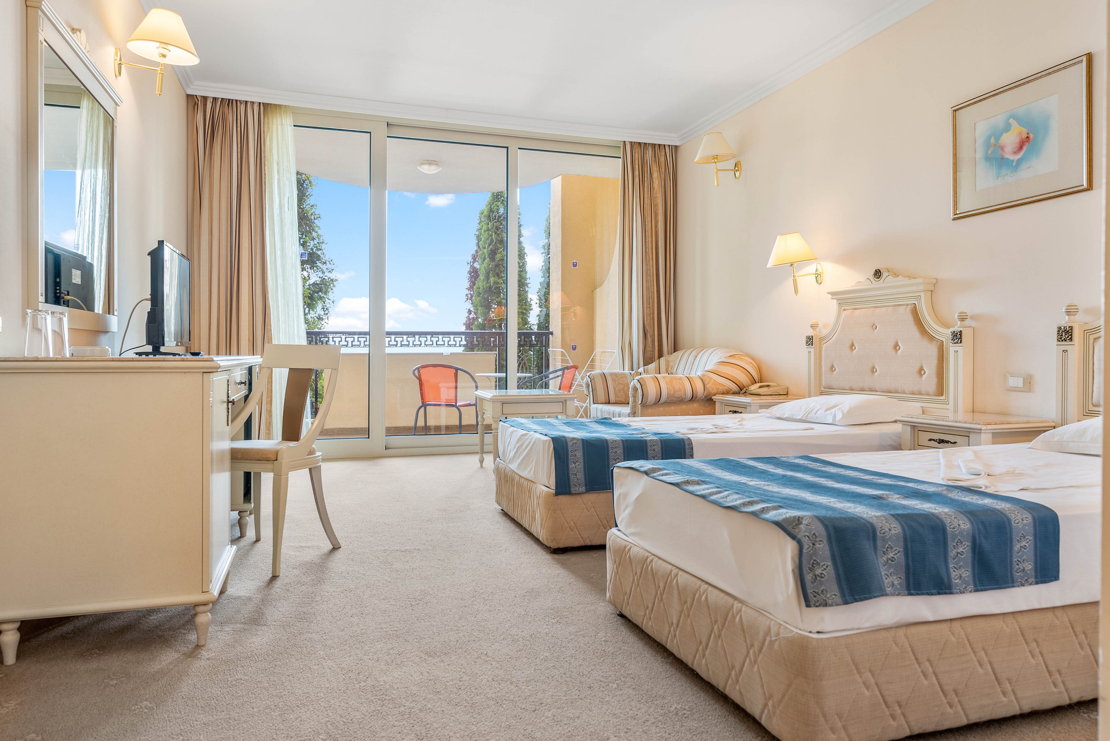 Well-lit hotel room featuring two neatly made twin beds with blue striped runners. A desk with a chair, television, and glassware sits to the left, while a cushioned armchair and side table add comfort. Large sliding glass doors open to a balcony with outdoor chairs and a view of trees and blue sky. Soft beige tones, white furniture, and warm lighting create a clean, inviting atmosphere.
