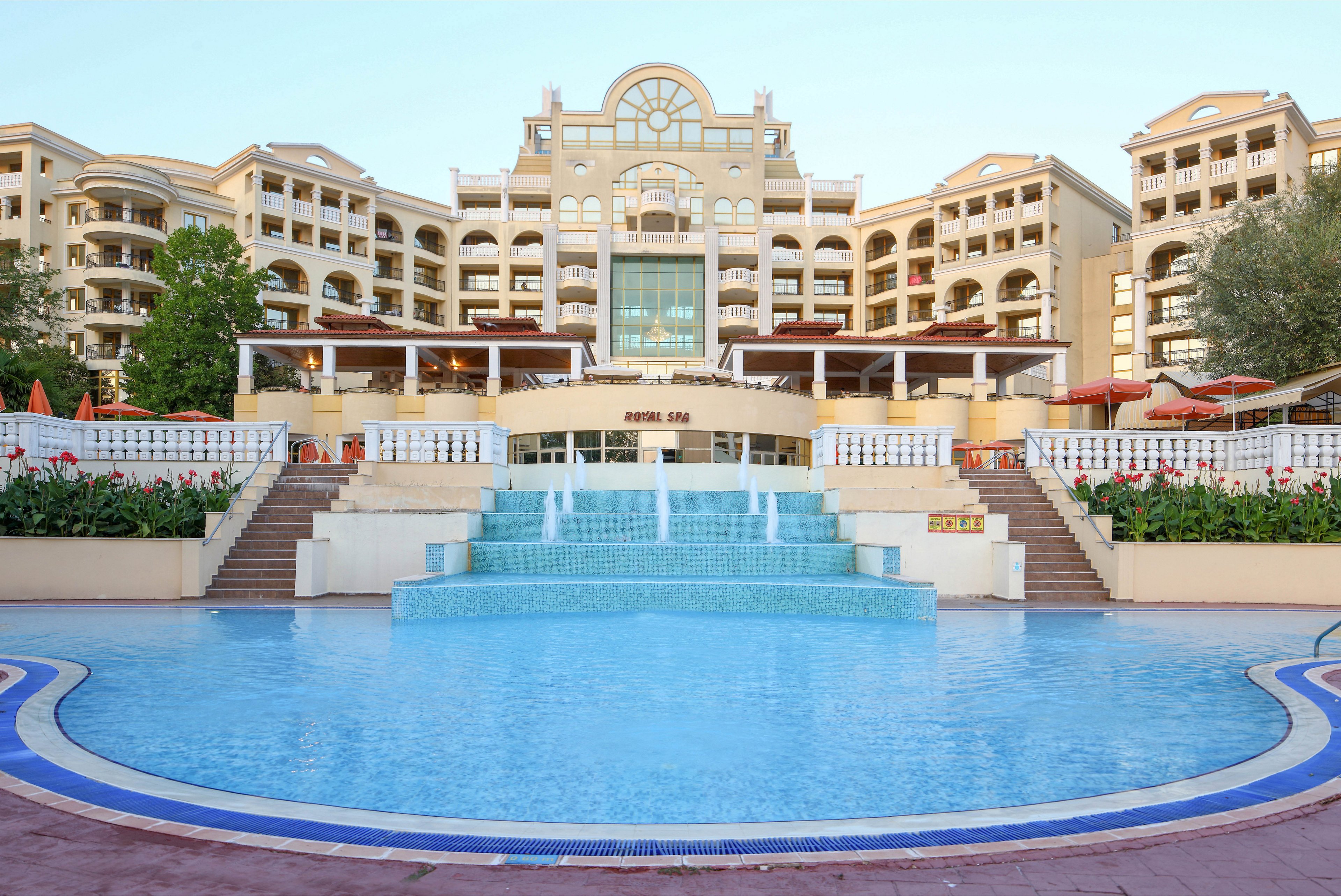 A grand resort hotel rises behind a large circular swimming pool with a tiered fountain at its center. The multi-story building features balconies, arched windows, and a symmetrical façade in warm beige tones. White railings, red flowers, and landscaped greenery frame the pool area, creating an inviting vacation atmosphere. The scene is captured in soft daylight under a clear blue sky, emphasizing the tranquil, upscale setting.