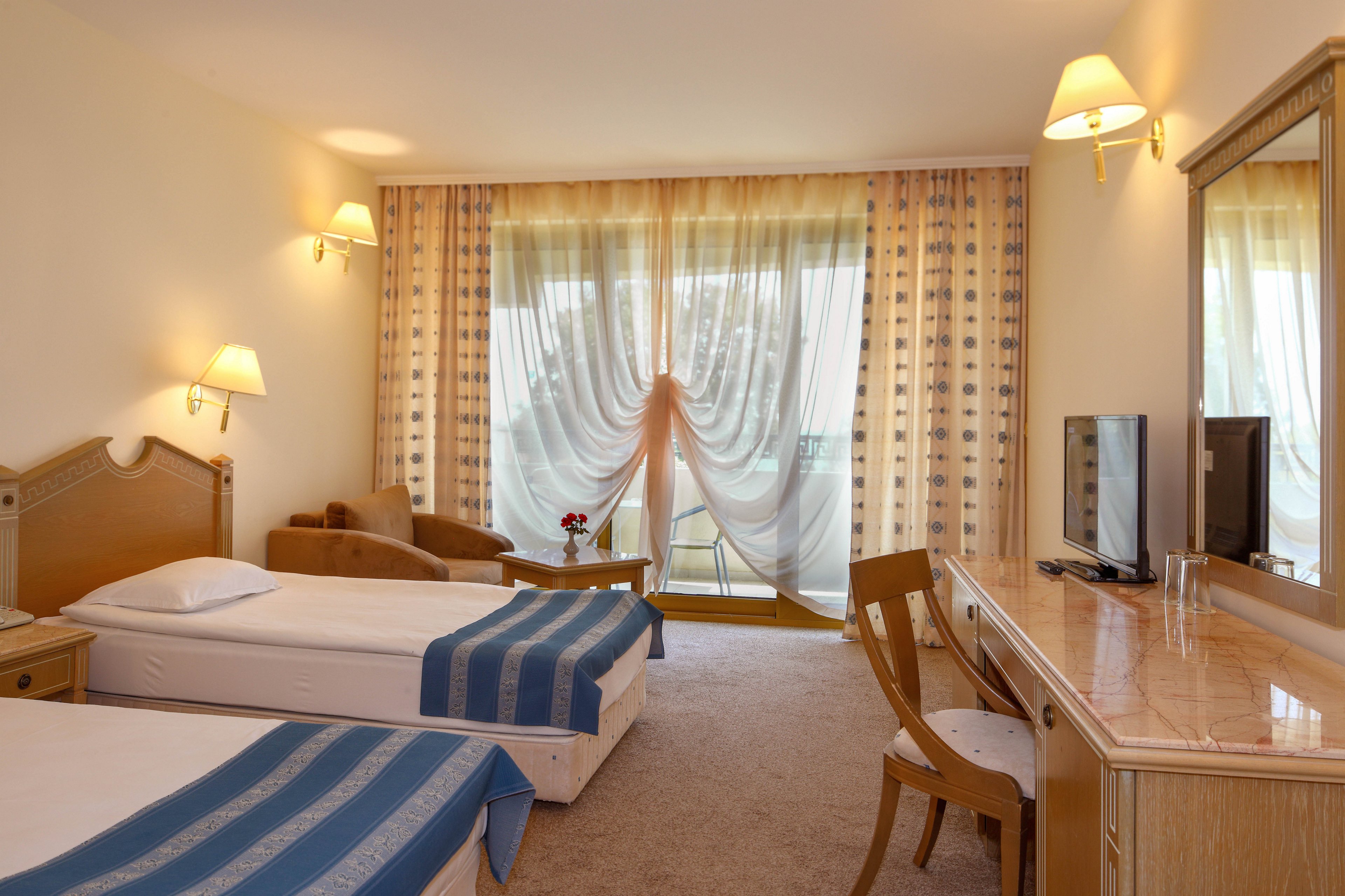 Well-lit hotel room featuring two single beds with white linens and blue striped runners. A wooden desk with a chair faces a flat-screen television and glassware on the right side. Sheer curtains and patterned drapes cover large balcony doors, revealing an outdoor seating area. Warm wall sconces and neutral beige tones create a cozy, inviting atmosphere.