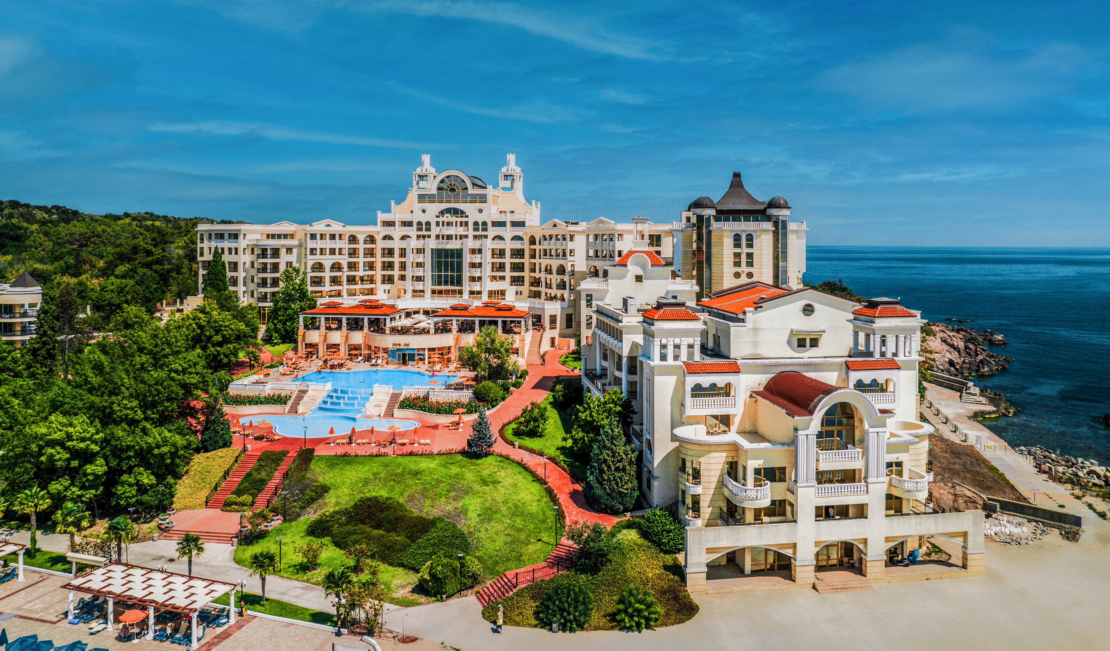 A large, ornate resort complex stands beside a calm blue sea, featuring multiple buildings with towers and decorative facades. In the center, a spacious outdoor pool area with sun loungers is surrounded by landscaped green lawns and red pathways. The foreground shows terraced walkways and garden beds leading toward the waterfront promenade. Clear skies and bright sunlight create a vibrant, inviting vacation atmosphere.