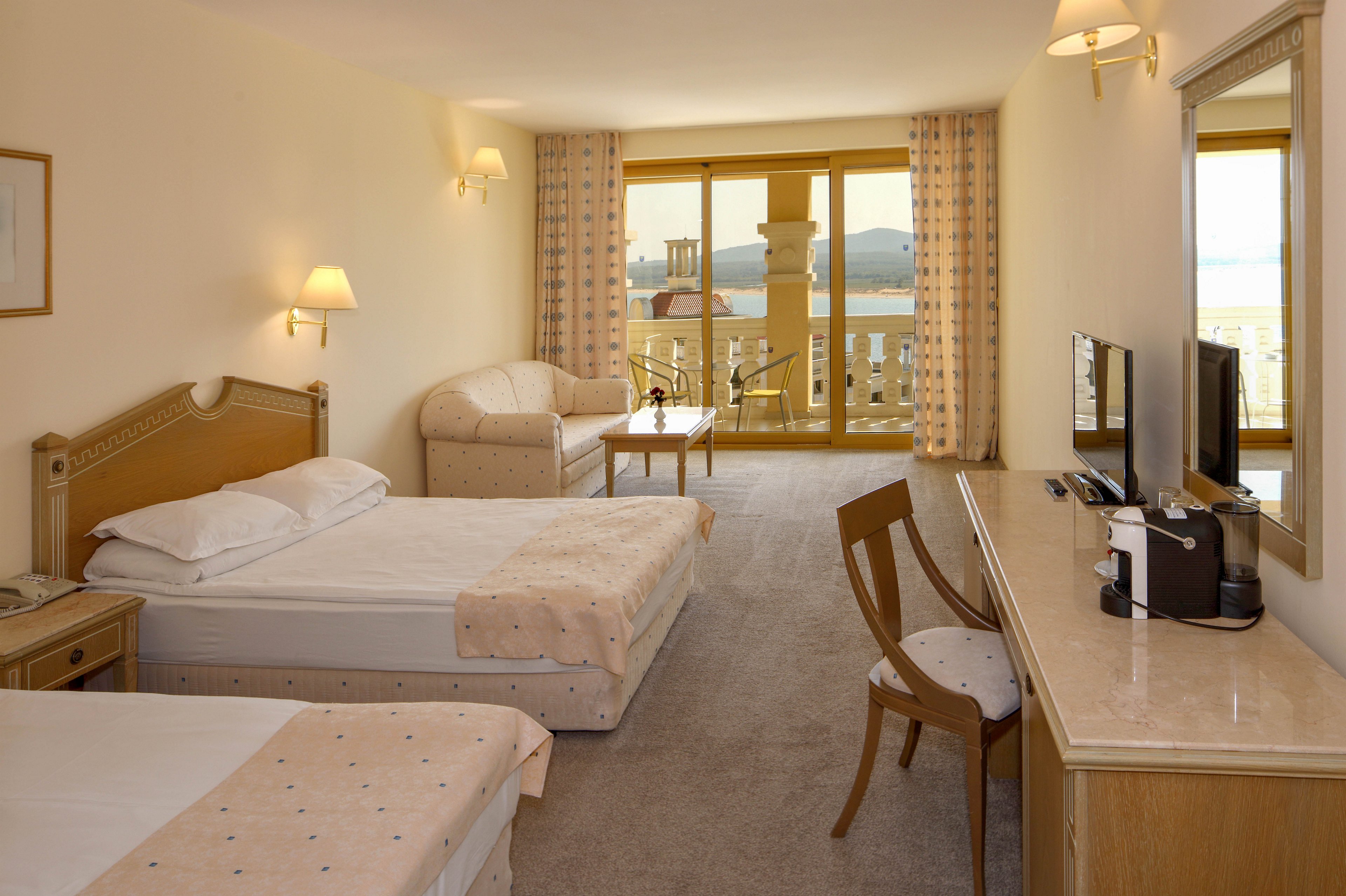 A spacious hotel room features two single beds with light-colored bedding and a matching sofa. A desk with a flat-screen television and wooden chair lines one wall, while wall-mounted lamps provide warm ambient lighting. Large glass doors open onto a balcony with outdoor furniture and a scenic view of the sea and distant hills. Neutral tones and soft patterns create a calm, inviting atmosphere suitable for relaxation or vacation stays.