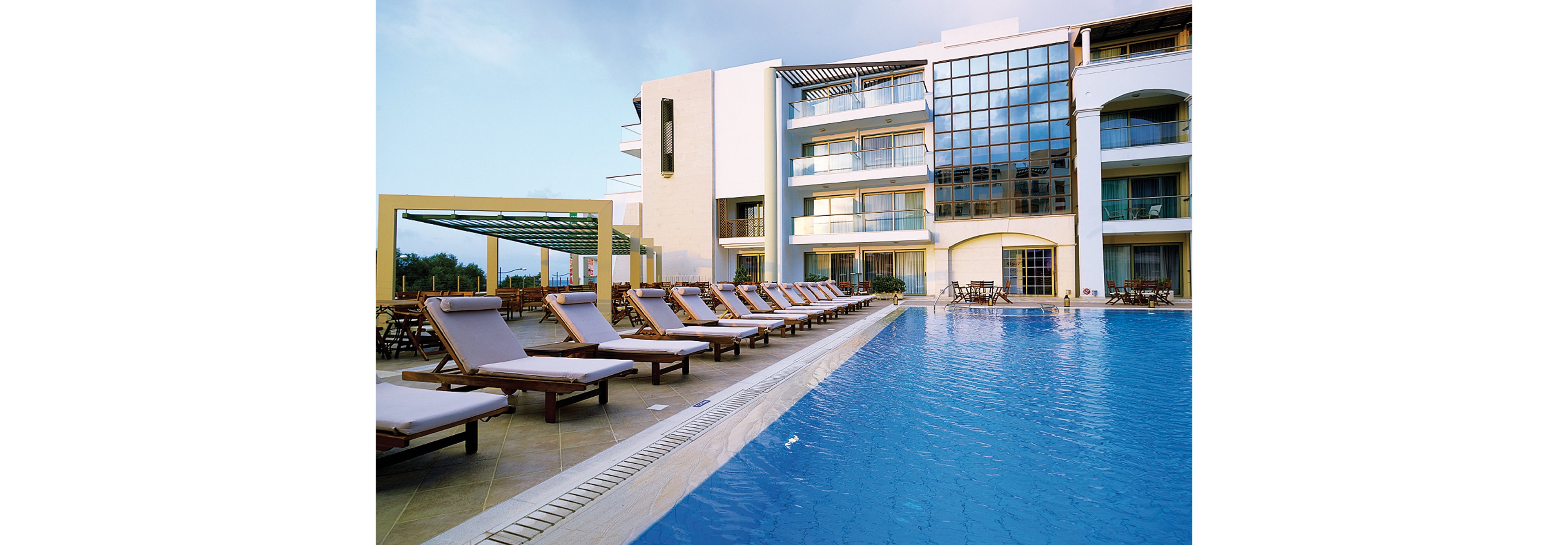 A serene outdoor pool area at a luxury resort featuring multiple lounge chairs arranged along the poolside. The setting includes modern architecture with large glass windows and balconies. The clear blue water of the pool reflects the sky, creating a tranquil atmosphere.