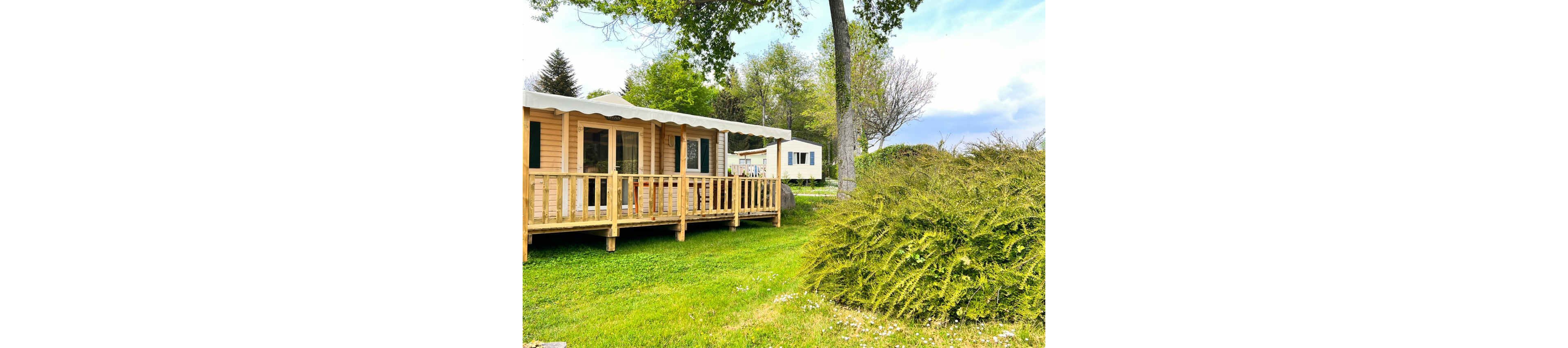 A wooden mobile home with a covered porch stands on a well-kept grassy plot. Another mobile home is visible in the background, suggesting a quiet campsite or holiday park. Tall trees and shrubs surround the area, adding greenery and shade. The scene feels peaceful and inviting, ideal for outdoor relaxation or vacation stays.