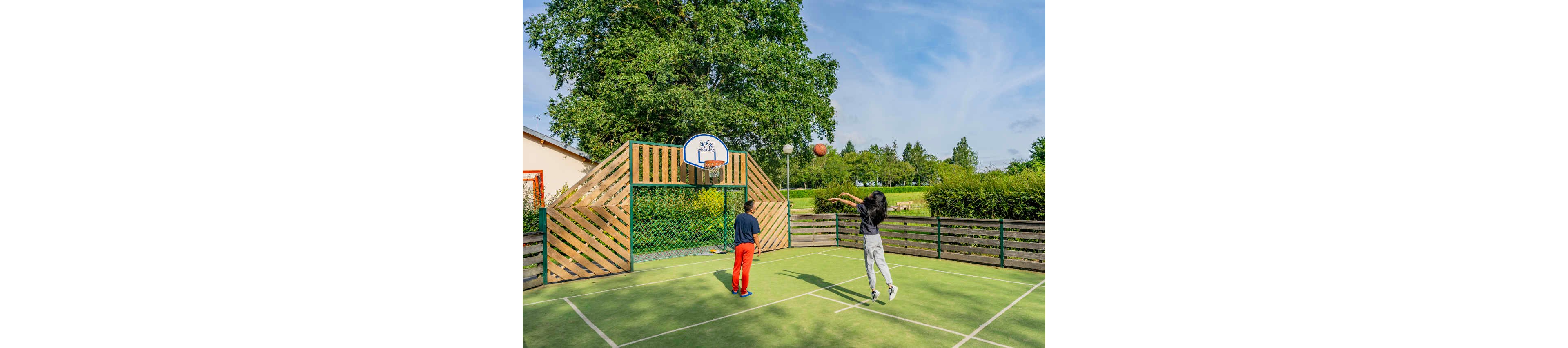 Two children play basketball on a small outdoor court surrounded by wooden fencing. One child shoots the ball toward the hoop while the other stands near the basket watching. The court is set in a green park-like area with trees, grass, and a clear sky in the background. The scene feels active and playful, capturing a casual sports moment in a sunny outdoor setting.