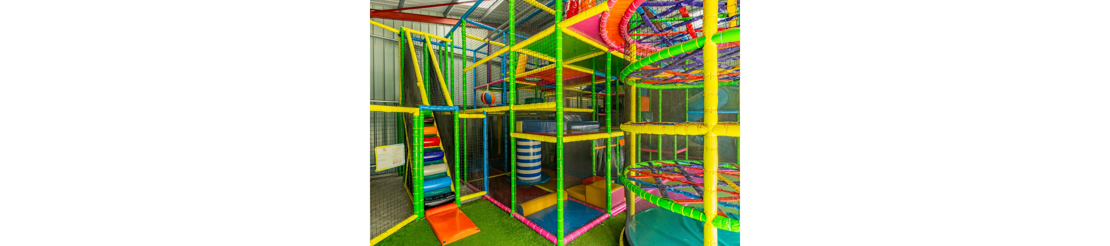 Brightly colored soft play equipment fills an indoor playground. The structure features padded climbing frames, slides, tunnels, and platforms enclosed with safety netting. Vibrant greens, yellows, blues, and reds create a fun and energetic atmosphere. Artificial grass flooring and cushioned surfaces emphasize safety for active children.
