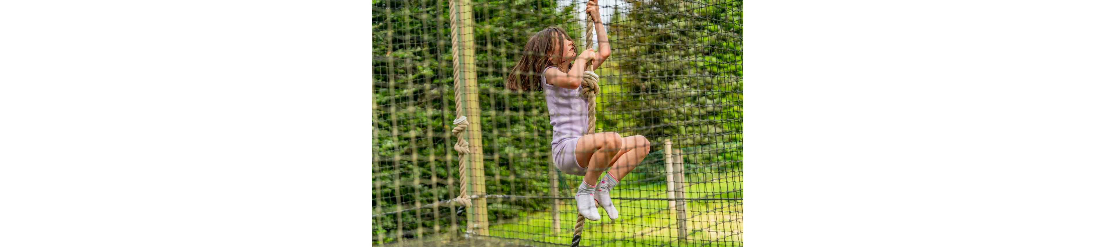 A young girl is captured mid-climb on a thick rope in an outdoor adventure park. She wears a light-colored outfit and socks while gripping the rope tightly with both hands and legs. The scene is framed by safety netting and wooden posts, with lush green trees in the background. The image conveys energy, concentration, and playful outdoor activity.