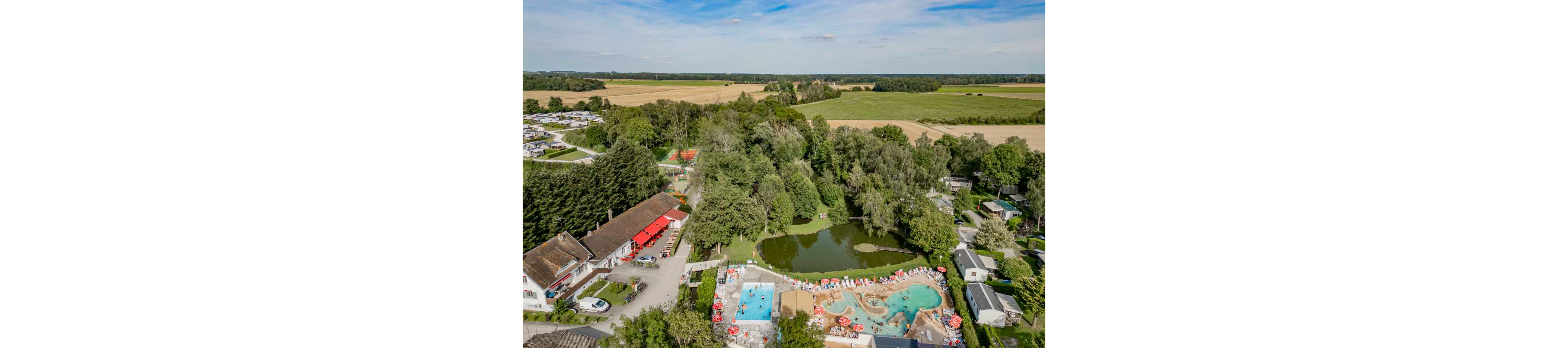 A large campsite is shown from above, surrounded by dense trees and open farmland. In the center, a natural pond and multiple outdoor swimming pools with sun loungers and umbrellas create a lively recreation area. Several buildings, caravans, and mobile homes are scattered around the site, connected by small roads and paths. The scene is bright and sunny, emphasizing the relaxed holiday atmosphere in a rural setting.