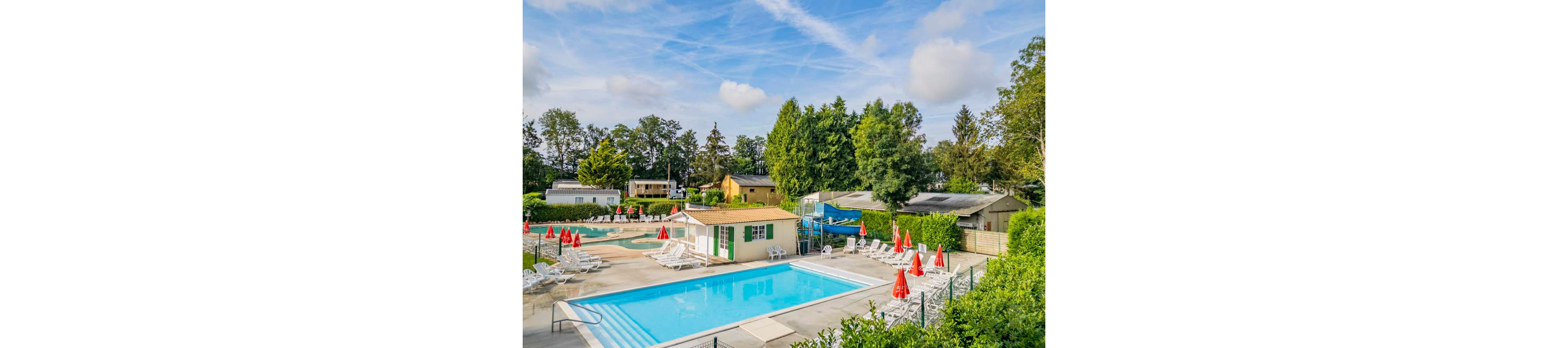 An outdoor swimming pool area is shown at a campsite or holiday park. A bright blue main pool and a smaller pool are surrounded by white sun loungers and red umbrellas. Small buildings and mobile homes sit among lush green trees in the background. The sky is partly cloudy with visible contrails, creating a relaxed vacation atmosphere.