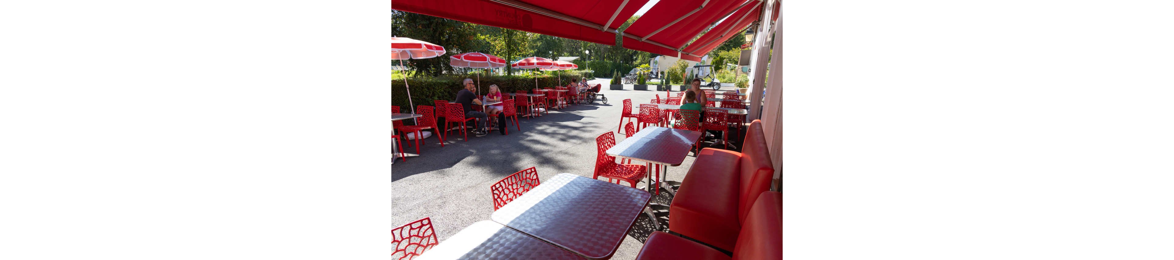 An outdoor café terrace features bright red chairs, benches, and awnings over metal tables. A few people sit and chat at the far end of the patio, enjoying the sunny weather. Red umbrellas line the edge of the seating area, with green trees and hedges in the background. The scene suggests a relaxed, casual atmosphere at a small street café.