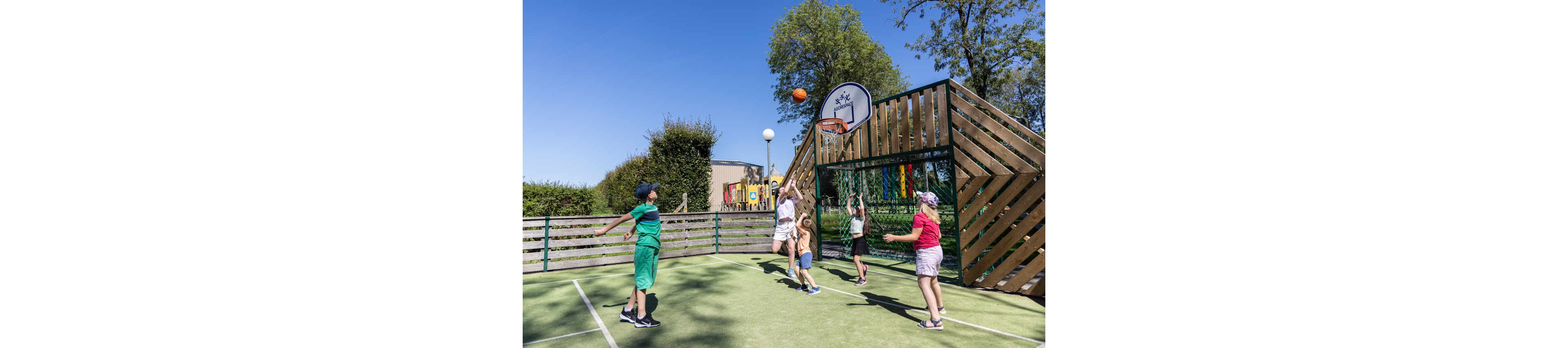 A group of children play basketball together on an outdoor court. One child jumps to shoot the ball toward the hoop while others watch and prepare to rebound. The court is surrounded by a wooden and metal fence structure with a mounted basketball backboard. Trees, greenery, and a clear blue sky create a bright, sunny atmosphere. The scene conveys active play, teamwork, and outdoor recreation.