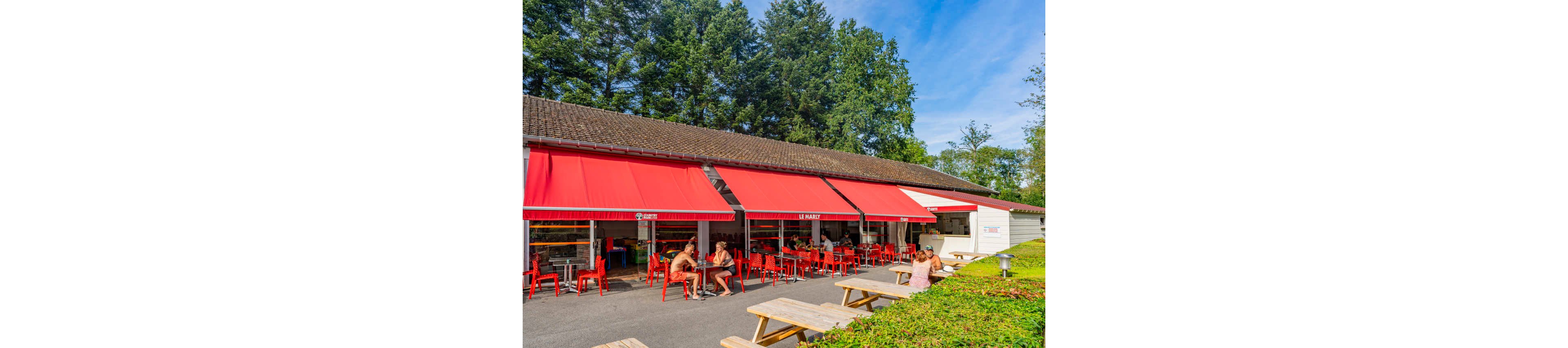 An outdoor café with bright red awnings and matching red chairs is set alongside a low building surrounded by tall green trees. People sit at tables enjoying food and drinks in the sunshine, creating a relaxed summer atmosphere. Wooden picnic tables are placed in the foreground near a neatly trimmed hedge, extending the seating area. The scene suggests a casual dining spot in a park or recreational area.
