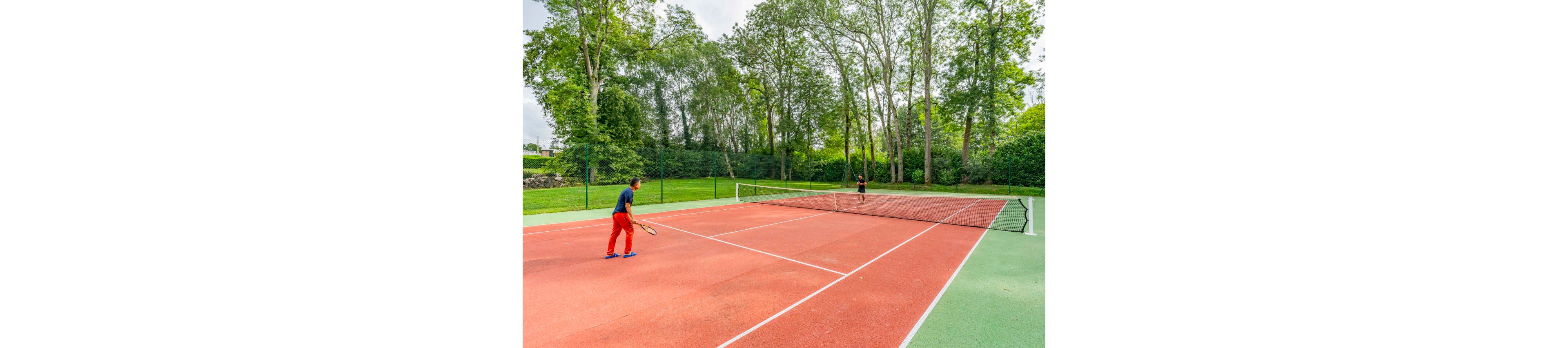 Two people are playing a game of tennis on a large outdoor court. The red hard court is surrounded by a tall green fence and lush trees. One player prepares to return the ball while the other stands near the opposite baseline. The scene suggests an active, recreational match in a bright, natural setting.