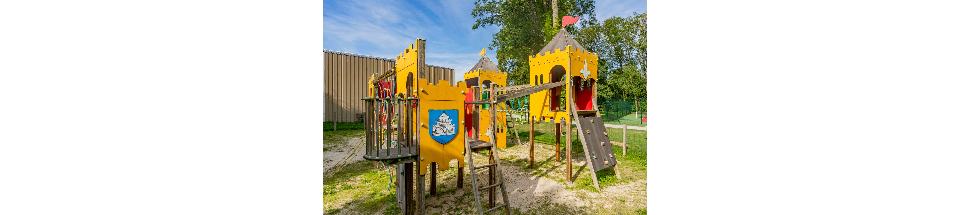 A bright yellow castle-themed playground structure stands in an outdoor park. The playset features towers, slides, ladders, and bridges designed to resemble a medieval fortress. A blue shield with a white castle illustration decorates one of the panels. The area is surrounded by grass, sand, trees, and nearby fencing, suggesting a safe recreational space for children.
