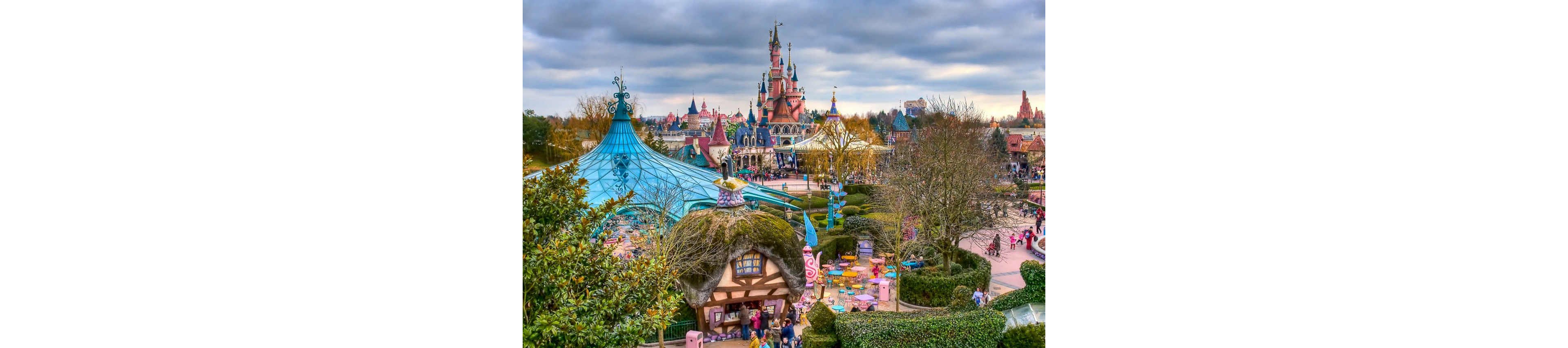 A vibrant fairytale theme park features a central pink castle rising above whimsical buildings and attractions. Colorful tents, teacup rides, and winding paths are surrounded by manicured greenery and trees. Visitors stroll through the park, adding scale and liveliness to the scene. A dramatic cloudy sky contrasts with the bright, playful colors of the attractions.
