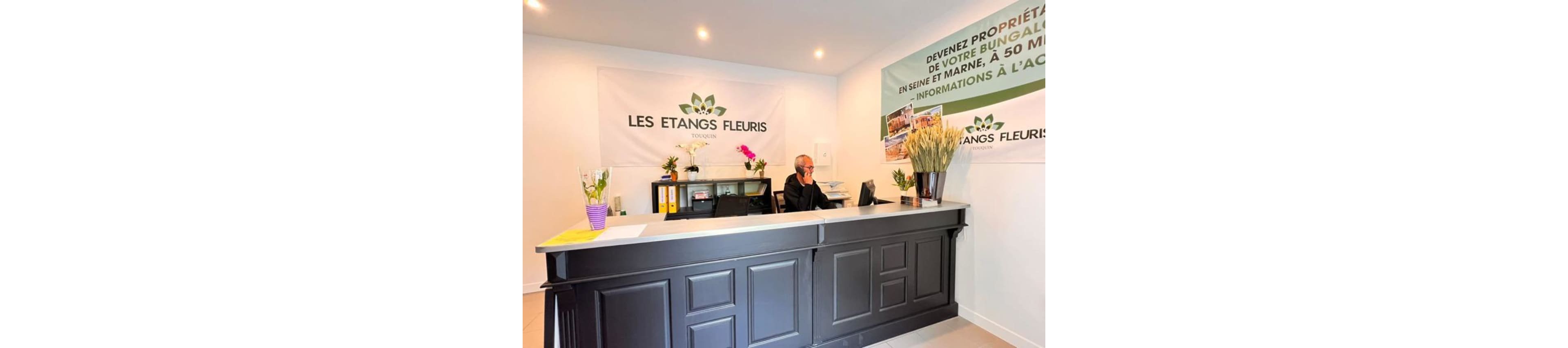 A receptionist sits behind a dark front desk in a bright office for Les Etangs Fleuris. The person is on the phone, with folders, a computer, and office supplies arranged on the counter. Large wall banners display the Les Etangs Fleuris name and a promotional message about property and bungalows. Potted plants and flowers add color and a welcoming atmosphere to the interior space.