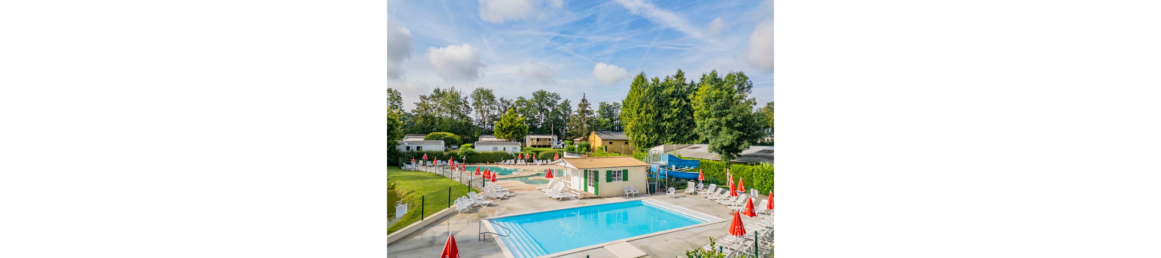 An outdoor swimming pool complex is set within a green holiday resort. White lounge chairs and closed red umbrellas surround the clear blue water. A small building with green shutters stands beside the main pool, with an additional shallow pool and water slide visible in the background. Trees, hedges, and scattered mobile homes frame the scene under a bright, partly cloudy sky.