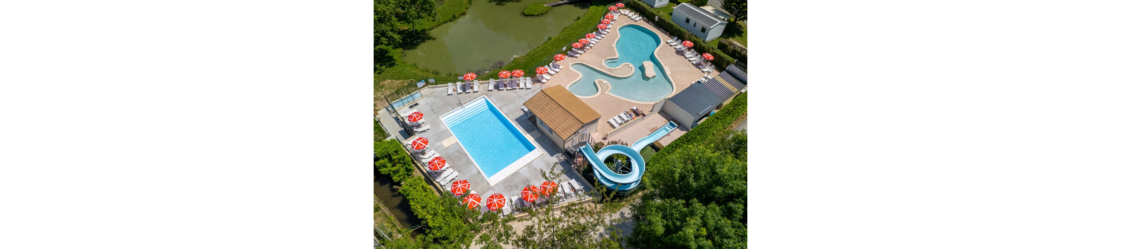 A resort-style outdoor swimming pool complex is captured from above on a sunny day. The scene features a rectangular lap pool, a curvy shallow pool, and a blue waterslide beside a central service building. Red and white umbrellas line the pool decks, with numerous white lounge chairs arranged neatly around the water. The complex is surrounded by greenery, a small pond, and nearby mobile homes or cabins, creating a relaxed vacation atmosphere.