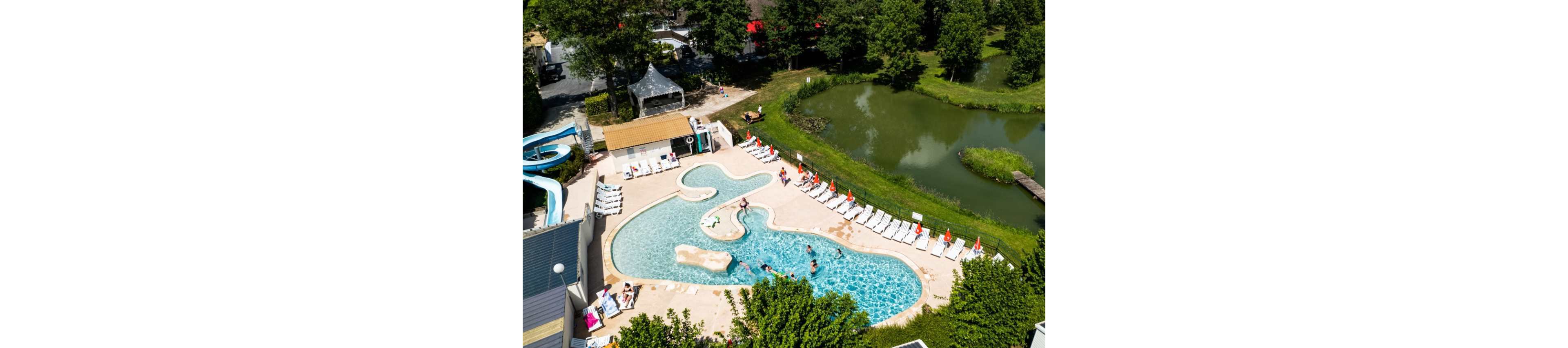A curving outdoor swimming pool is seen from above at a holiday resort. People relax on white loungers lined with red umbrellas around the pool while others swim and play in the water. A small waterslide and nearby buildings are visible on one side, with a green pond and trees surrounding the area. The scene suggests a sunny summer day in a family-friendly vacation setting.
