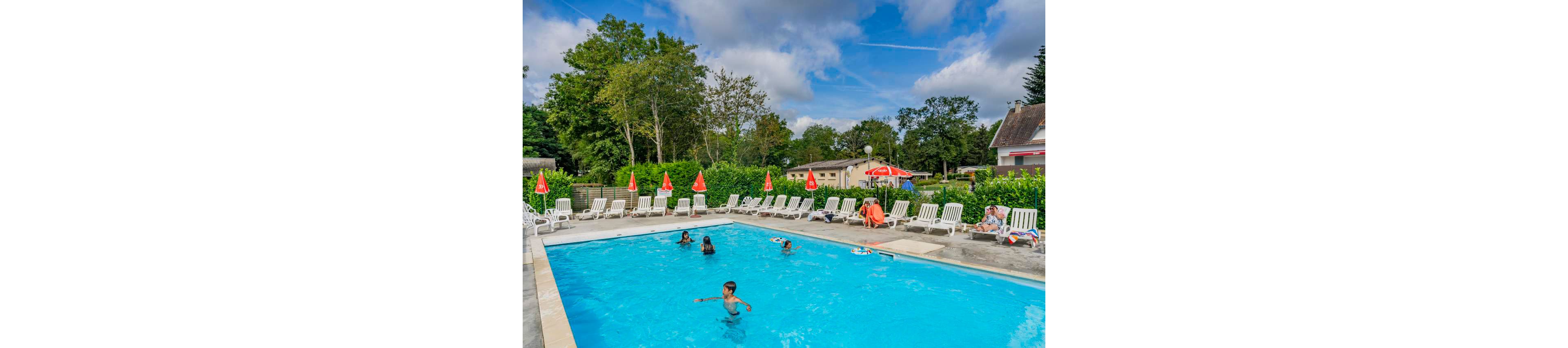 An outdoor swimming pool is surrounded by white lounge chairs and red umbrellas at a holiday resort. Several children and adults enjoy swimming and relaxing by the clear blue water. Green trees and hedges frame the pool area, with low buildings and houses visible in the background. The scene is bright and sunny with scattered clouds in a blue sky, creating a cheerful vacation atmosphere.