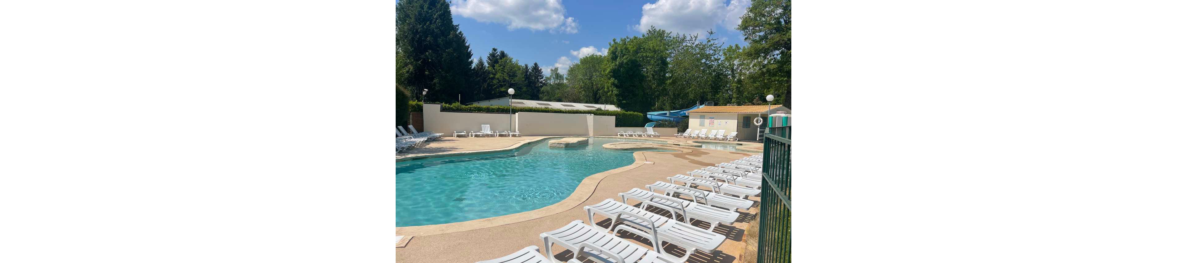 An outdoor swimming pool is surrounded by numerous white lounge chairs under a bright, sunny sky. The pool area appears calm and empty, with clear blue water and a curved pool shape. In the background, there are tall green trees, a small building, and a blue water slide. The overall scene suggests a quiet resort or campground pool ready for visitors.