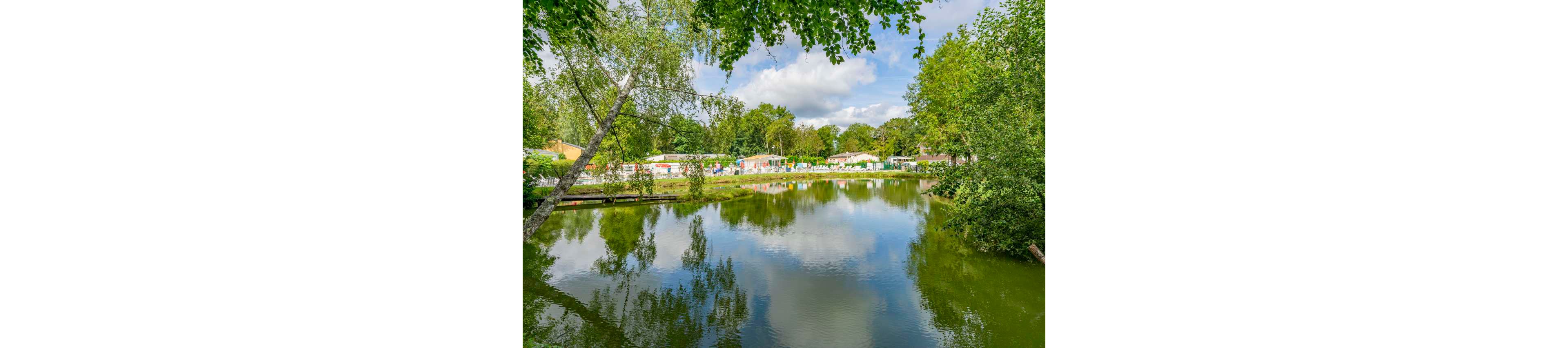 A calm lake reflects surrounding trees and a bright blue sky with scattered clouds. On the far shore, a row of caravans and holiday homes is visible among the greenery. The scene suggests a peaceful vacation park set in a wooded area. Lush foliage frames the water, emphasizing the natural, tranquil atmosphere.