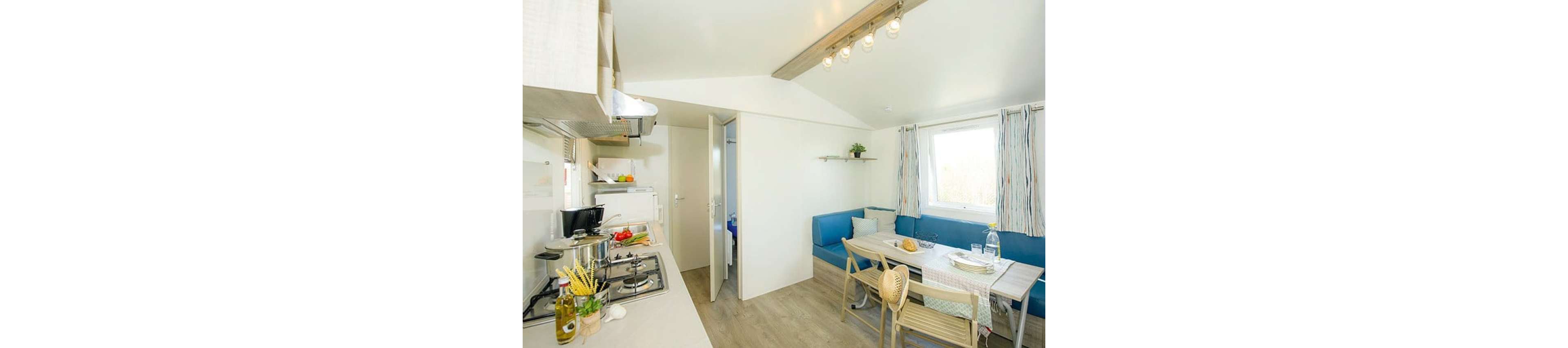 A small modern kitchen opens into a cozy dining and seating area in a bright room. The space features a gas stove with pots and fresh vegetables prepared on the counter. A wooden dining table is set with chairs, a bench, and a checked tablecloth near a large window with striped curtains. Light wood flooring and white walls create an airy, minimalist atmosphere with simple decor.