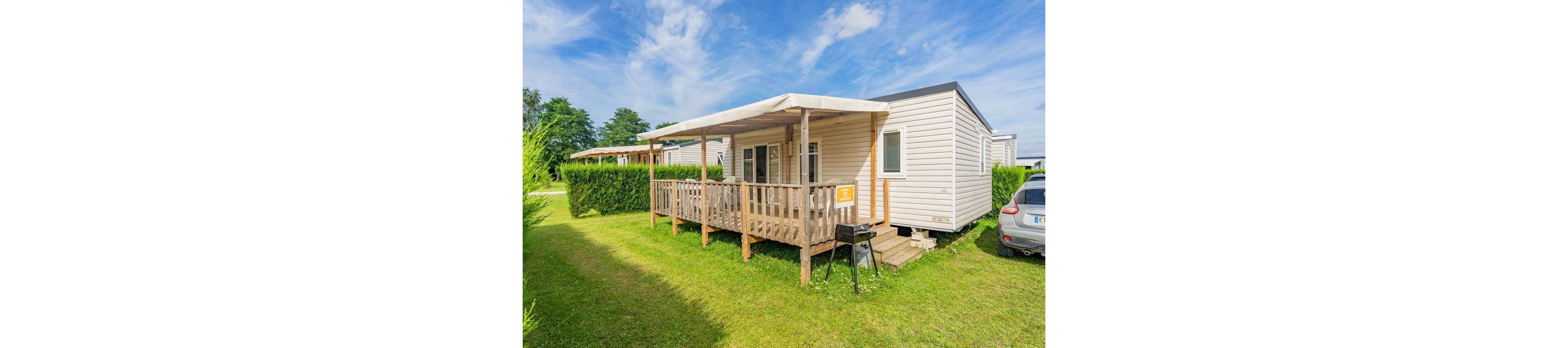A light-colored mobile home with a covered wooden terrace stands on a neatly mowed grassy plot. The setting appears to be a quiet campsite or holiday park with hedges and other units in the background. A small charcoal grill and outdoor table are placed near the steps leading to the terrace. A parked car is partially visible to the right, suggesting a vacation or weekend getaway atmosphere under a bright blue sky with scattered clouds.