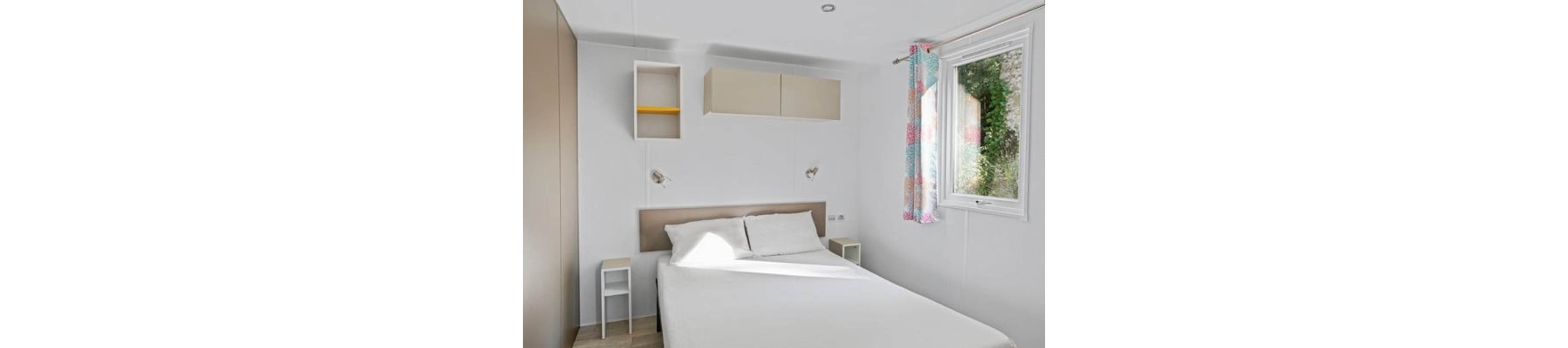 A small, bright bedroom features a neatly made double bed with white linens against a light-colored headboard. Two compact bedside tables flank the bed, each with a wall-mounted reading light above. Simple wall cabinets and an open shelf provide storage on the white wall. A window with colorful patterned curtains lets in natural light and offers a view of greenery outside, creating an airy and inviting atmosphere.