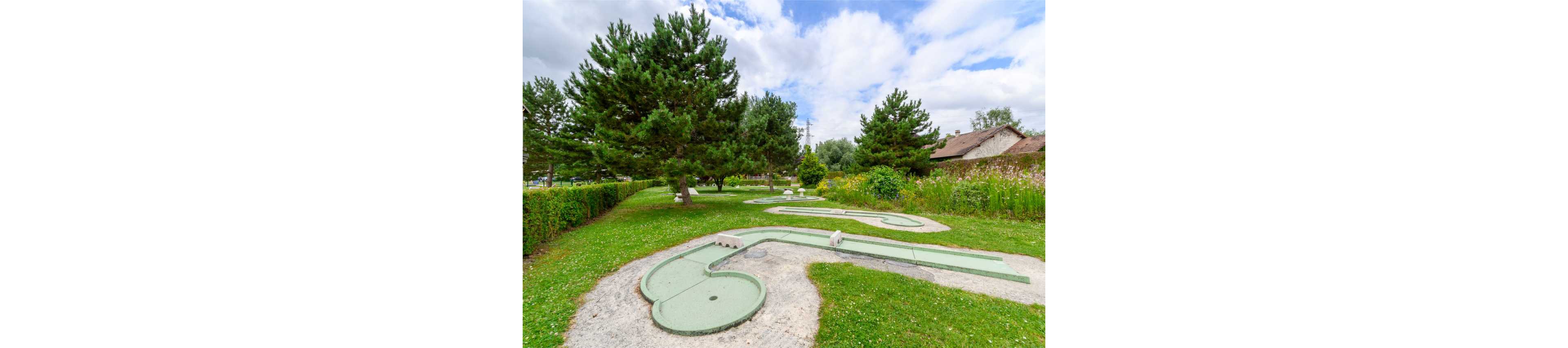 An outdoor mini golf course winds through a lush green park. Curved putting lanes with concrete borders are surrounded by neatly cut grass and tall evergreen trees. A hedge and wildflower area frame the course, with a house visible in the background. The scene is bright and calm under a partly cloudy sky, suggesting a relaxing recreational setting.