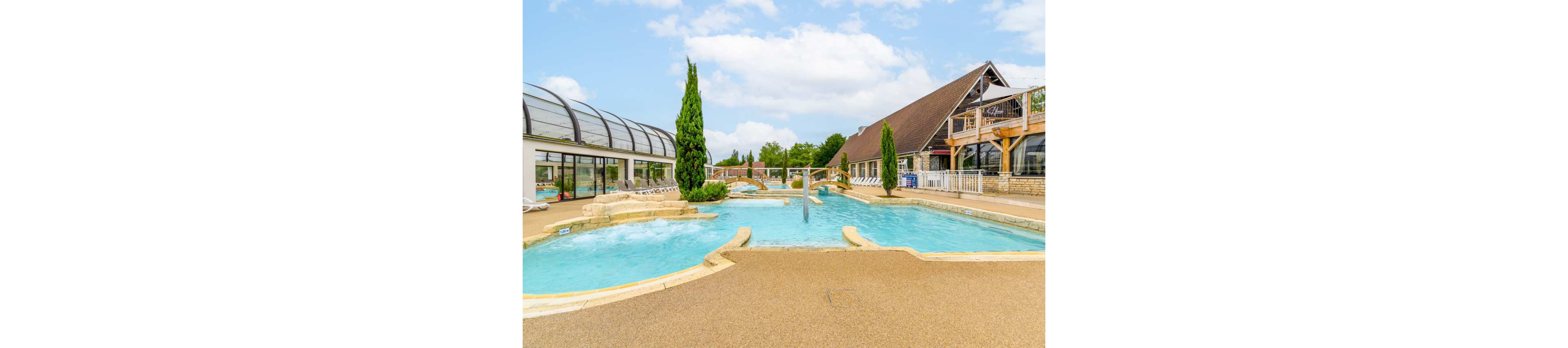 An outdoor swimming pool complex is shown at a leisure resort. The pool features curved edges, built-in seating, and water jets creating gentle bubbles. A covered glass structure with lounge chairs lines one side, while a rustic building with wooden beams and balconies stands on the other. Tall evergreen trees and a bright sky complete the relaxed holiday atmosphere.