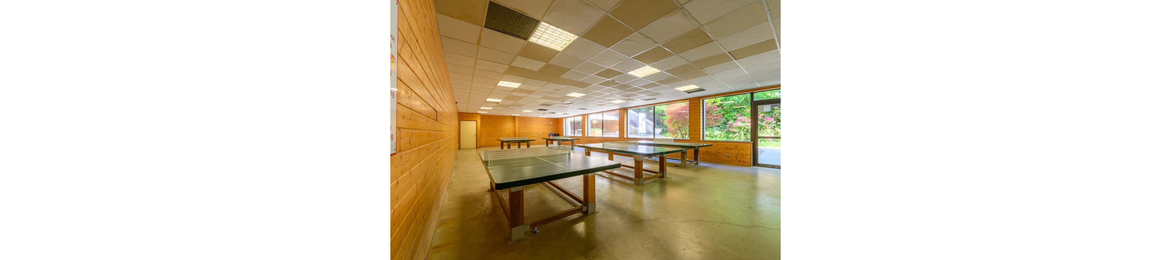 A large indoor recreation room features several green table tennis tables arranged in rows. The space has wood-paneled walls, a tiled drop ceiling, and polished concrete floors. Floor-to-ceiling windows along one side reveal lush greenery and outdoor steps. Fluorescent ceiling lights and natural daylight create a bright, inviting atmosphere suitable for games and group activities.