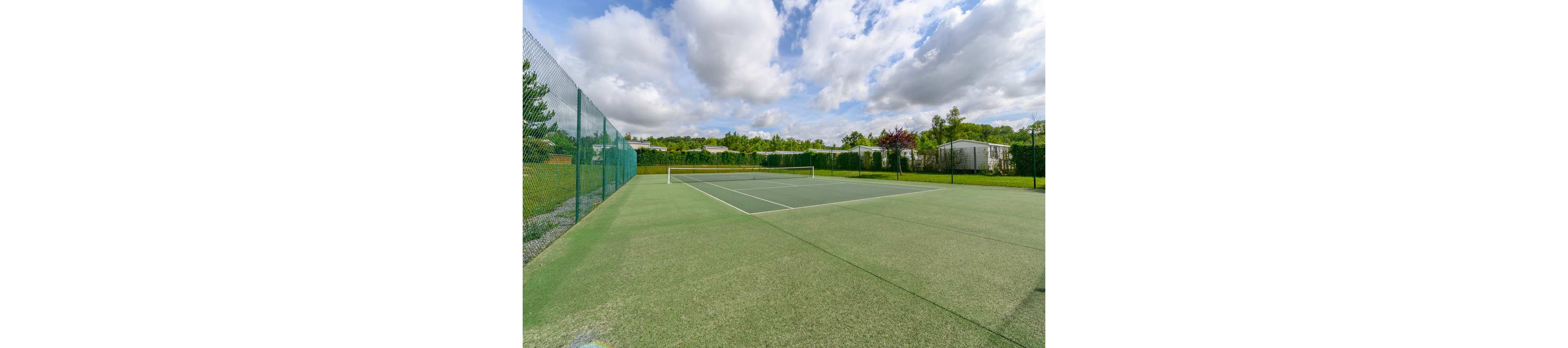 An empty green tennis court is shown under a partly cloudy sky. The court is surrounded by tall chain-link fencing and lush green hedges. In the background, several light-colored mobile homes or holiday cabins are visible among trees. The scene feels calm and spacious, suggesting a quiet recreational area.