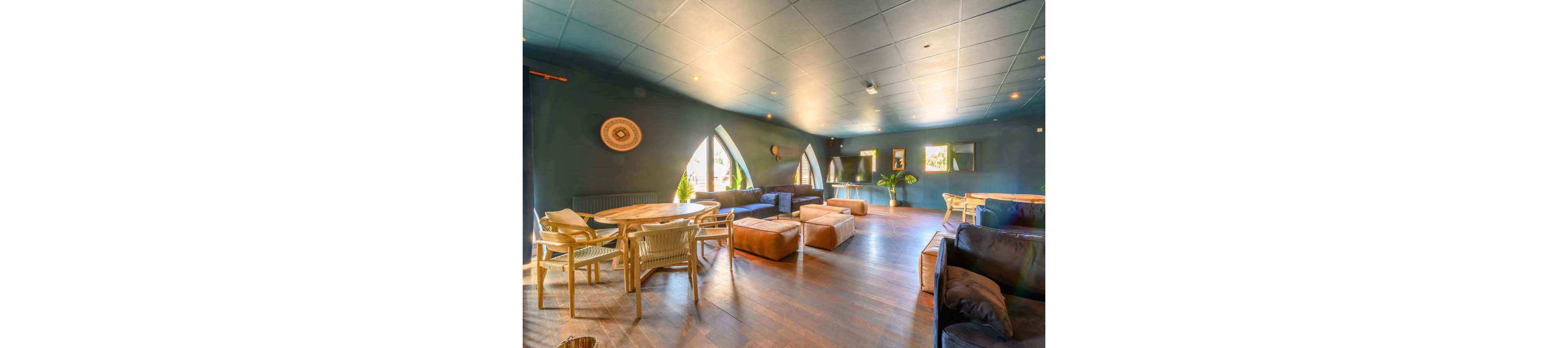 Spacious modern lounge interior featuring a mix of sofas, armchairs, and round wooden tables. Large arched windows allow natural light to brighten the dark blue walls and wooden floor. Soft ottomans and woven chairs create a relaxed, informal seating arrangement. Warm ambient lighting and decorative wall accents contribute to a cozy, welcoming atmosphere.