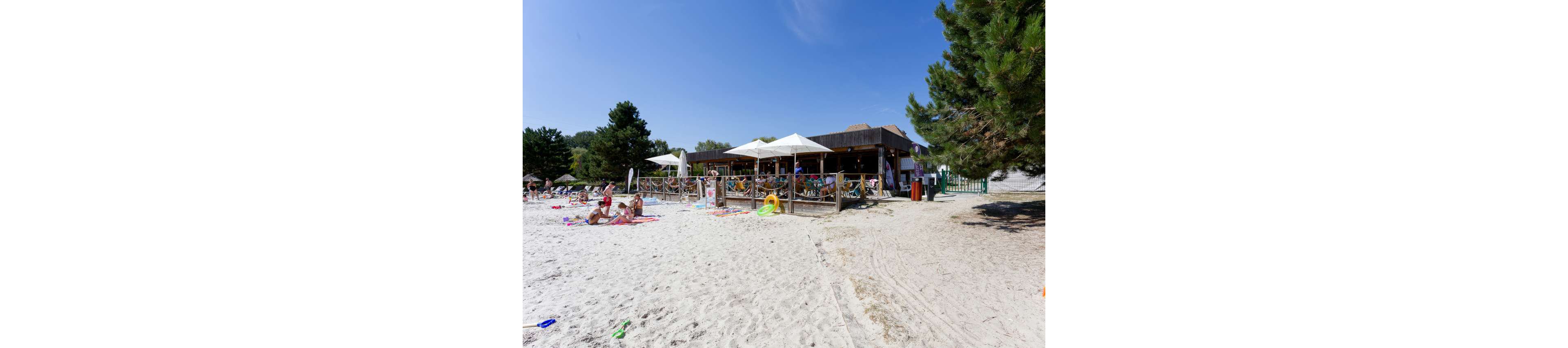 A wooden beach bar with large white umbrellas overlooks a sandy shoreline on a sunny day. Several people relax on towels and sunbathe near the water, while others sit in the shaded seating area of the bar. Green trees frame the scene, contrasting with the bright blue sky and pale sand. The overall atmosphere is relaxed and summery, suggesting a casual vacation setting.