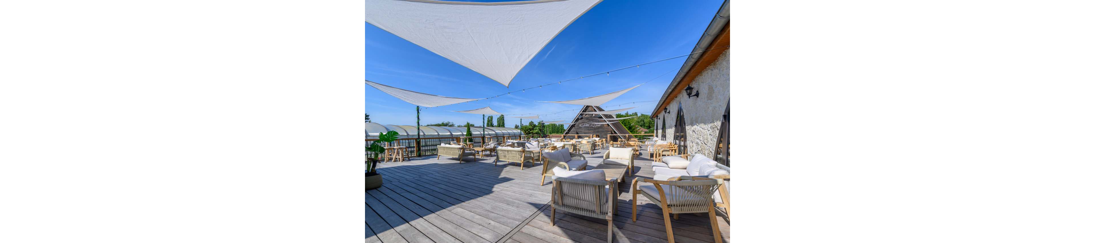 An open-air rooftop terrace features modern wicker lounge chairs and sofas arranged on a wide wooden deck. White triangular shade sails are stretched overhead, creating geometric patterns against a bright blue sky. The terrace overlooks greenery and a glass-covered structure in the background, suggesting a scenic outdoor venue. String lights add a festive touch, making the space suitable for relaxing, socializing, or hosting events in a calm, contemporary setting.