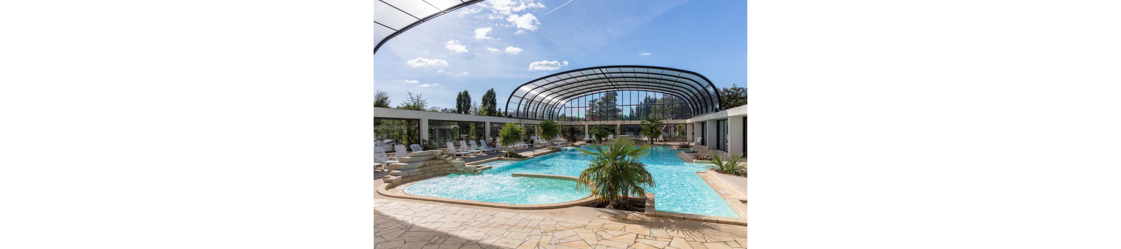 A large resort-style swimming pool sits beneath a modern curved glass roof structure. Sun loungers line the stone pool deck, suggesting a relaxing vacation setting. Palm trees and landscaped plants add a tropical feel around the turquoise water. The bright blue sky and scattered clouds enhance the sunny, inviting atmosphere.
