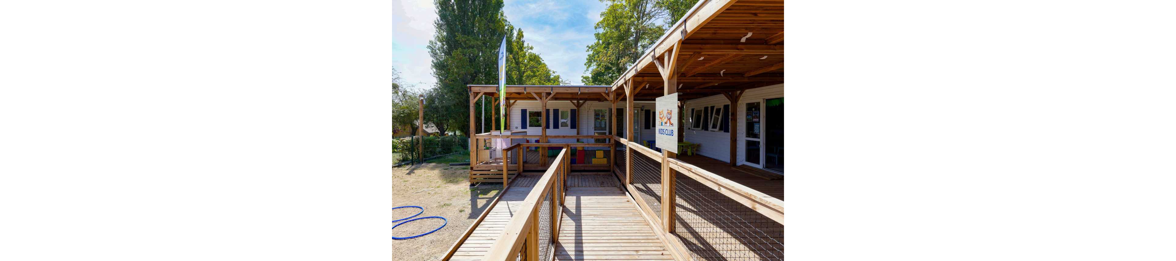 An outdoor kids club building features a spacious wooden deck and ramp leading to the entrance. The structure is surrounded by trees and open grounds, suggesting a campsite or recreational setting. Colorful play blocks and a sign reading "KIDS CLUB" with cartoon characters indicate a child-friendly environment. Natural light and wooden materials create a warm, welcoming atmosphere for children’s activities.