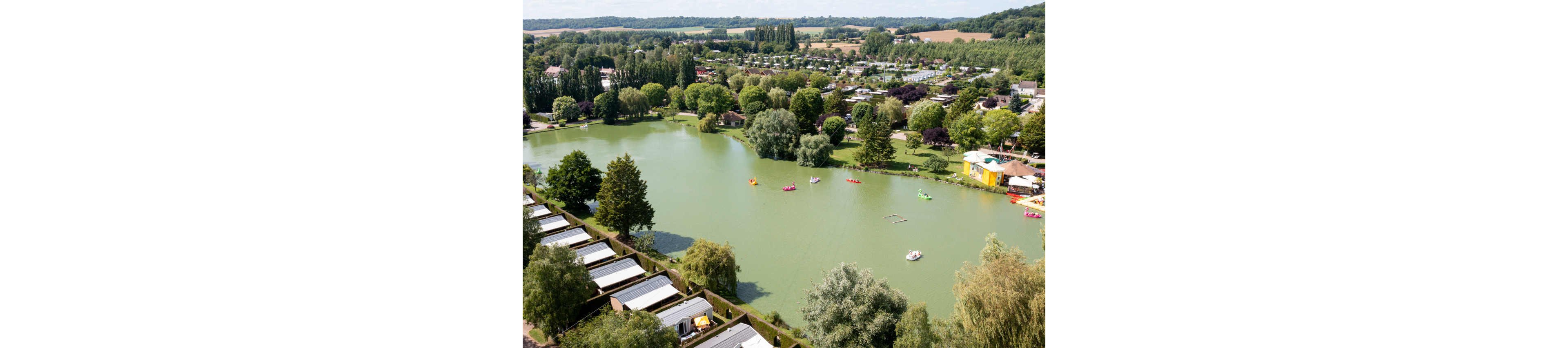 A large greenish lake is surrounded by a holiday park with rows of cabins and lush trees. Colorful pedal boats and small watercraft are scattered across the calm water. On the right side, a yellow building and waterfront area stand out among the greenery. Rolling fields and distant hills frame the background, creating a peaceful countryside setting.