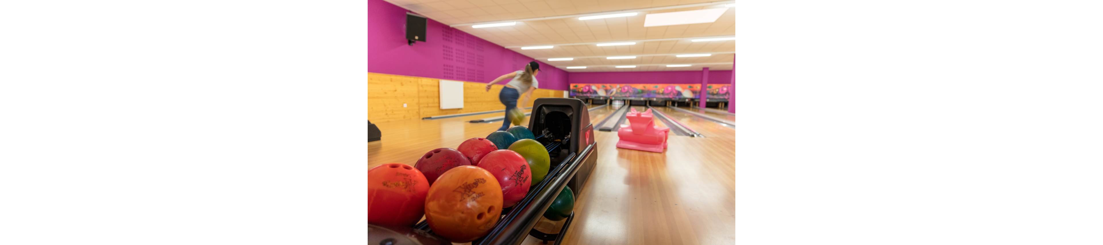 A row of brightly colored bowling balls sits in the ball return at a modern bowling alley. In the background, a person is captured in motion as they release a ball down the lane. The lanes are framed by a wooden floor, pink walls, and vibrant wall art, creating a playful atmosphere. Soft overhead lighting and a shallow depth of field emphasize the action and colorful equipment.
