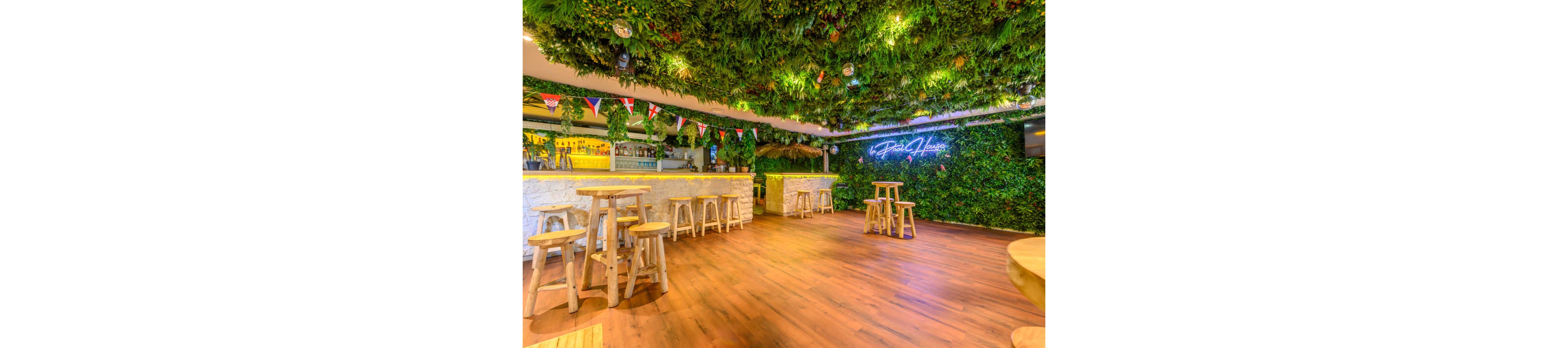 A bright tropical-themed indoor bar features wooden stools and high tables on a warm-toned wooden floor. The ceiling and walls are covered with dense green foliage, creating a lush, immersive atmosphere. Colorful bunting and hanging ornaments add a festive touch above the bar counters. A neon sign glows on the back wall, enhancing the vibrant, modern nightlife setting.