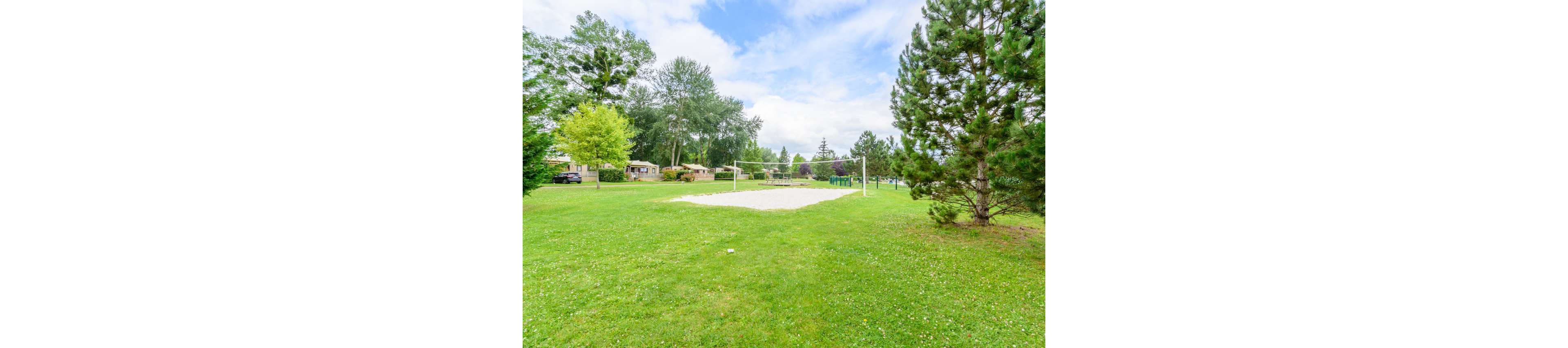 An outdoor sand volleyball court sits in the middle of a large grassy park. The court is surrounded by lush green trees and open lawn under a bright, partly cloudy sky. Residential houses are visible in the background, giving the scene a suburban neighborhood feel. The overall atmosphere is calm and inviting, ideal for recreation and community activities.