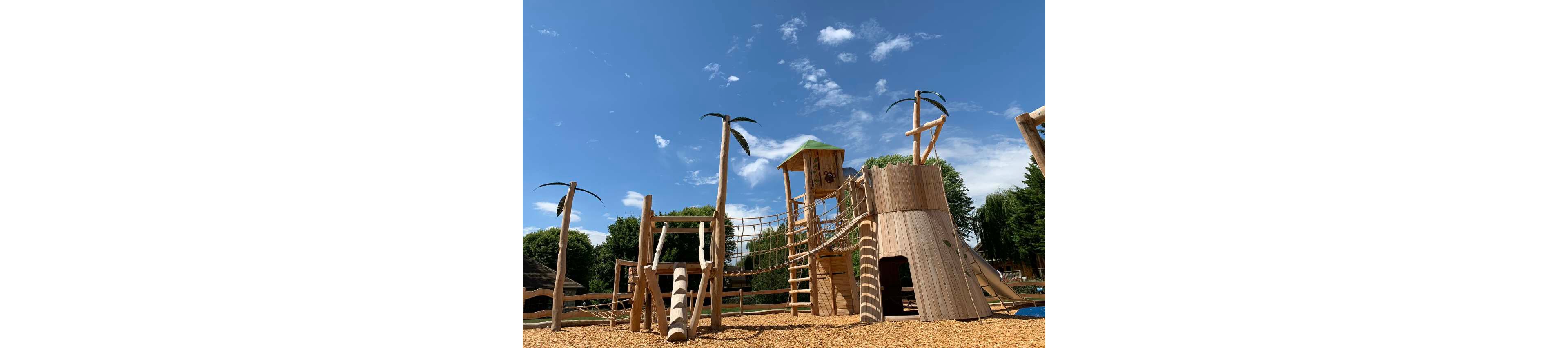 A large wooden adventure playground stands outdoors on a sunny day. The structure features towers, slides, climbing nets, and palm tree–shaped poles set on a bed of wood chips. Lush green trees surround the play area, contrasting with the vivid blue sky and scattered white clouds. The scene suggests an inviting, nature-inspired space for children’s outdoor play and exploration.