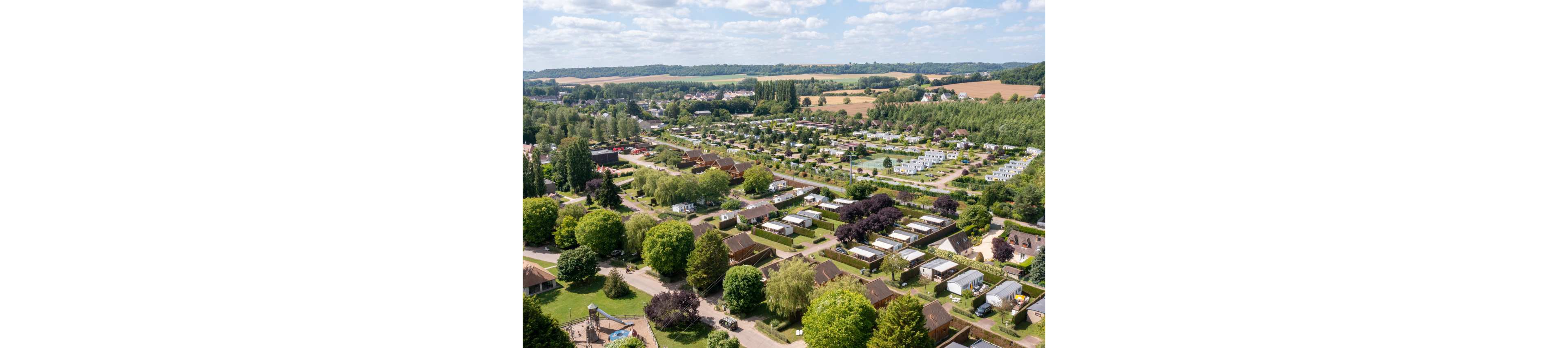 A wide aerial view shows a large caravan and chalet holiday park set in green countryside. Rows of white mobile homes and small cabins are arranged along tree-lined roads and grassy plots. Surrounding fields, distant hills, and scattered buildings indicate a rural village setting under a bright, partly cloudy sky. The lush trees and open spaces create a peaceful vacation atmosphere.