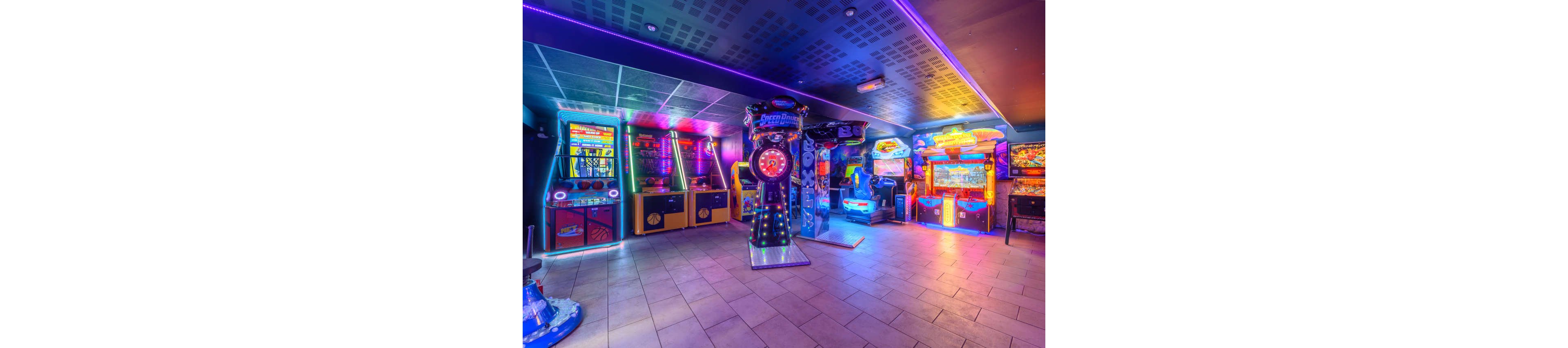 A brightly lit arcade room features multiple electronic game machines lined up against the walls. Neon and LED lights in blue, purple, and pink illuminate the tiled floor and ceiling. Various racing, ticket, and skill games stand ready for players, but the space is currently empty. The overall atmosphere feels energetic and playful, emphasizing the vivid colors and glowing screens.